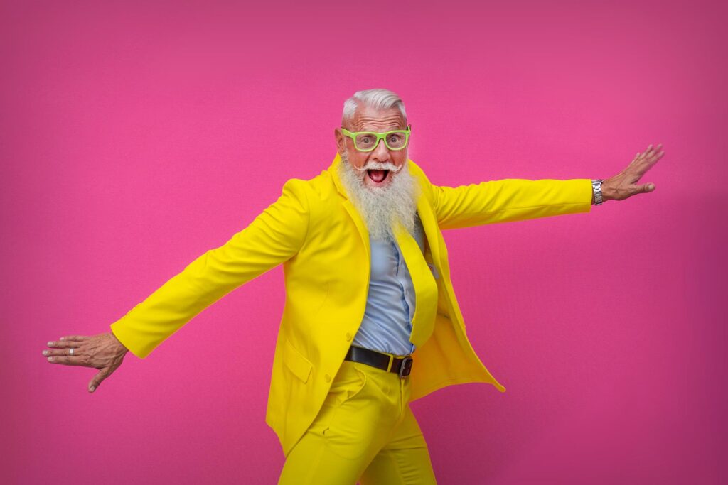 Elderly man with a long white beard, wearing a bright yellow suit and glasses, striking a playful pose against a pink background—a perfect fit for eye-catching advertising.