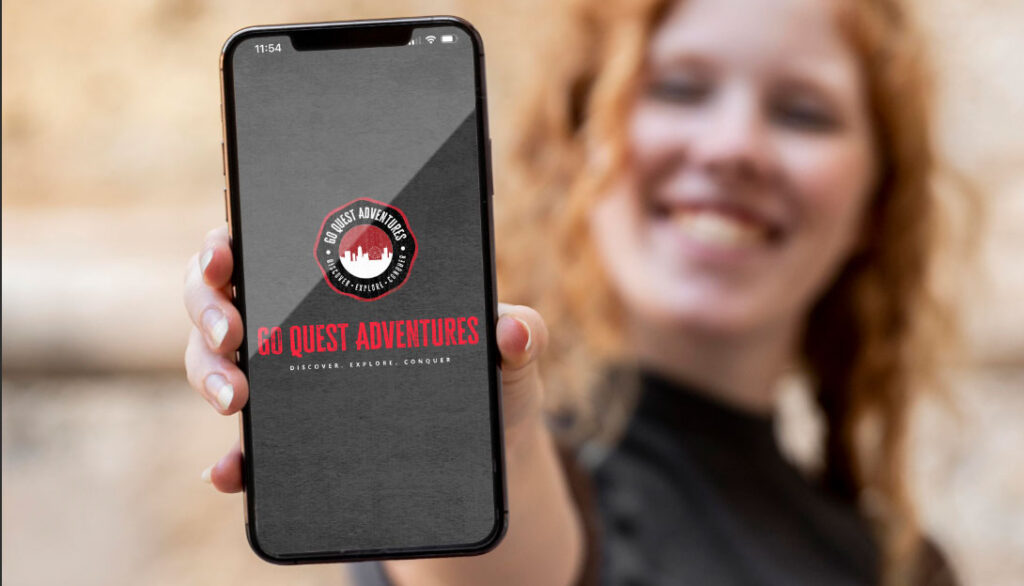 A person holds up a smartphone displaying the Go Quest Adventures app with its new logo on the screen, featuring the text, "DISCOVER. EXPLORE. CONQUER.