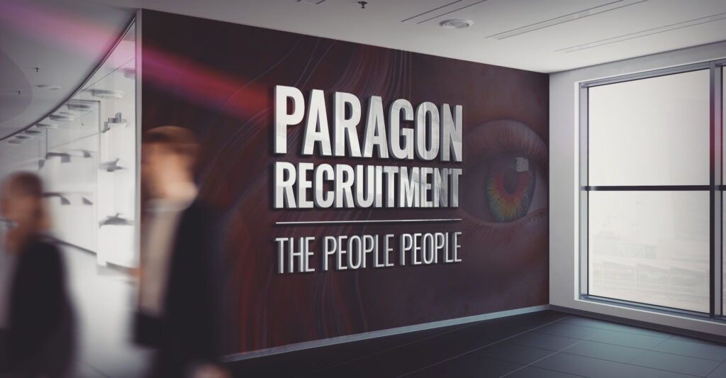 Blurry people walk past a wall with "Paragon Recruitment: The People People" written on it, emphasising their expertise in brand development, in a modern, bright office hallway.