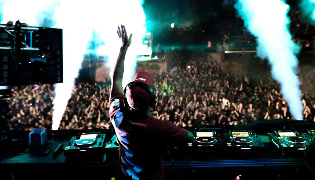 The DJ performs on stage with a large crowd, raising one hand amidst blue and white stage lights and smoke effects, creating a scene that's perfect for Social Media Design.