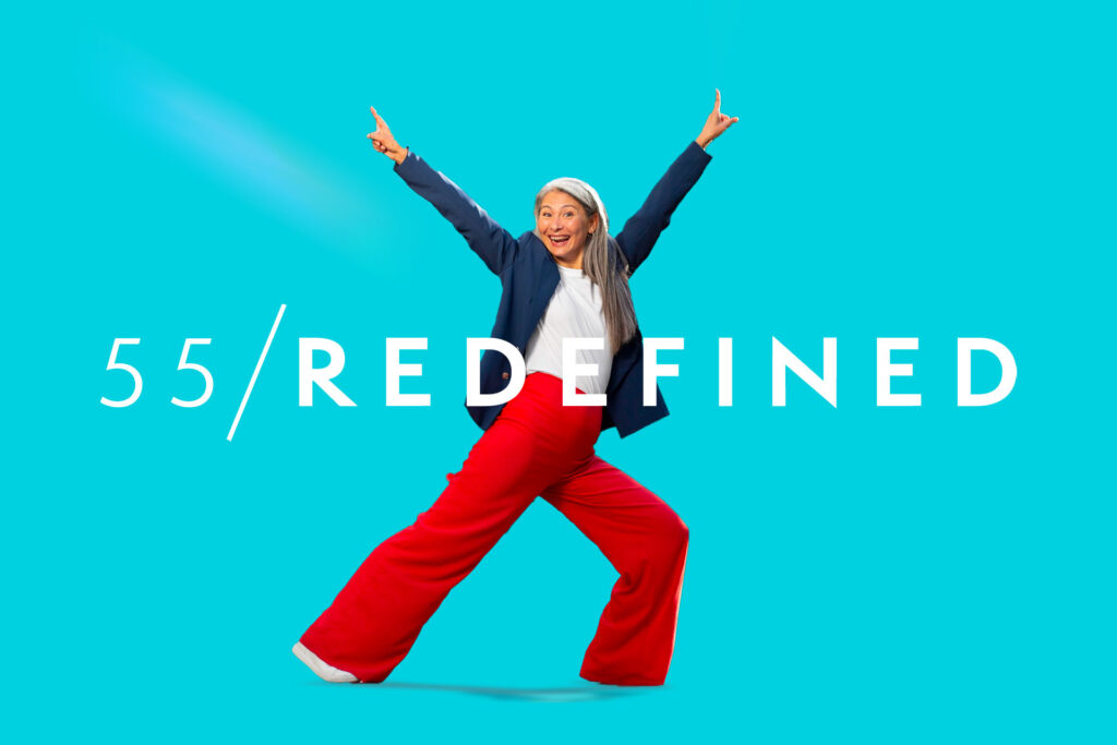 An older woman poses energetically with arms raised against a bright blue background. The text "55 / REDEFINED" is displayed across the image.